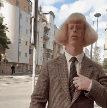 GIF d'un homme portant une perruque blonde coupée au bol et une moustache, en veste de tweed et cravate. Il se tient dans une rue et se désigne de la main d'un air sérieux.
